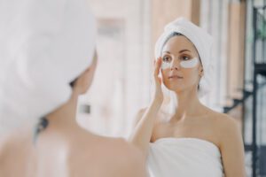 Rudeninė odos atgaiva: kaip po vasaros išgelbėti paakius ir susigrąžinti švytėjimą Moteris tepanti paakių kremą rudeninės šviesos fone, prižiūrinti odą po vasaros.